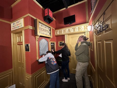 Three players work on puzzles in a red and gold theater lobby set next to a ticket booth
