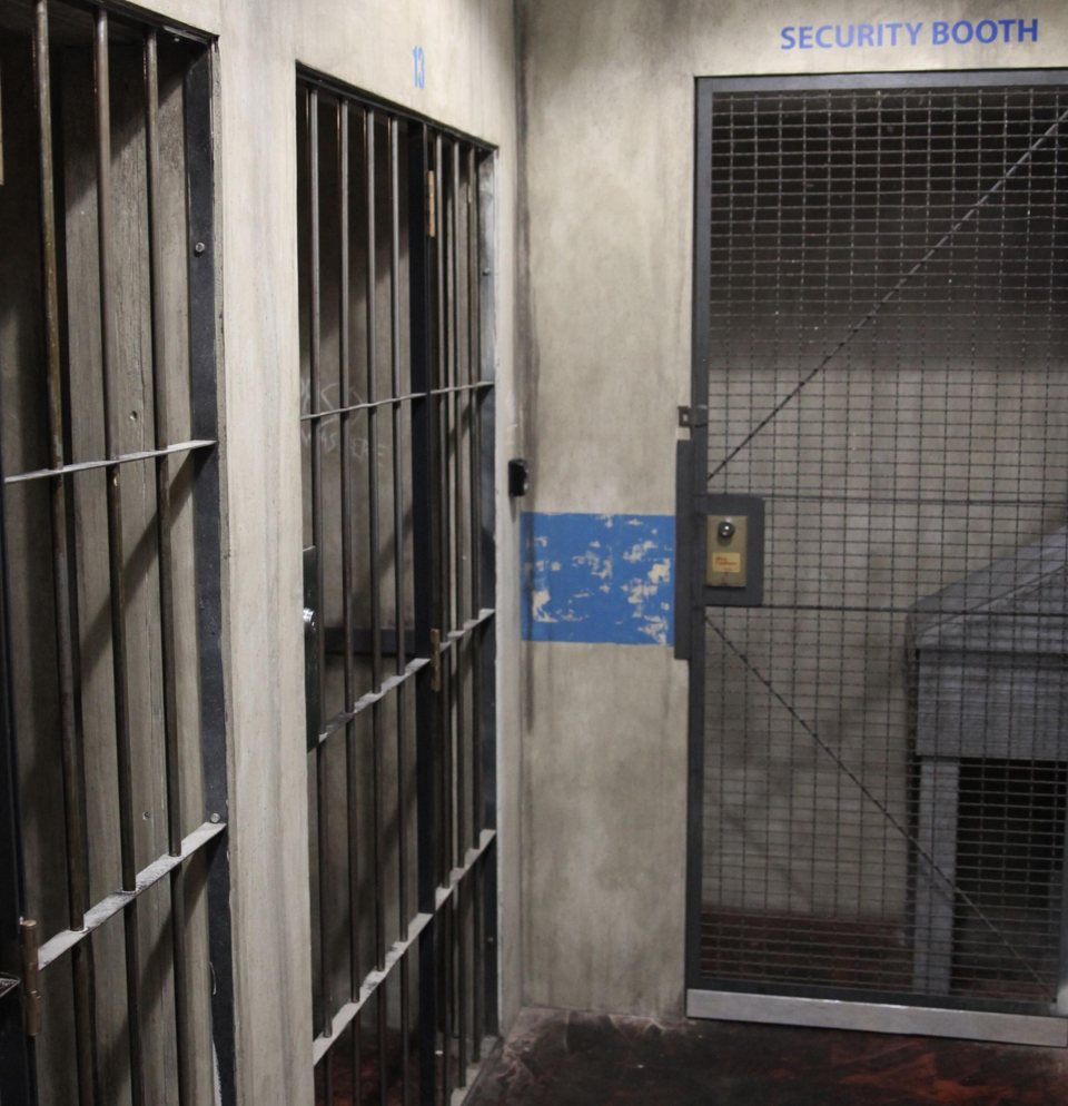 Barred jail cells and a wire-mesh security booth along a worn concrete corridor