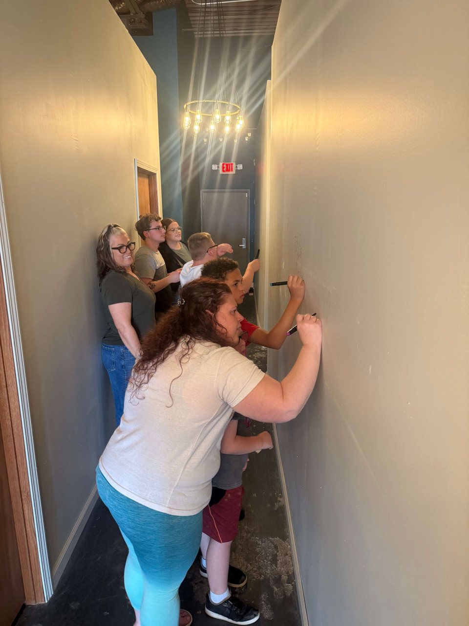 A team solves a puzzle with markers on the wall of a narrow, light corridor under a bare-bulb chandelier