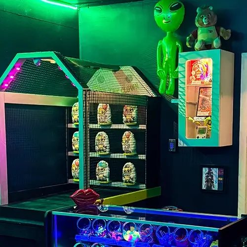Green neon-lit retro room with an inflatable alien, a caged shelf of capsules and a teddy bear