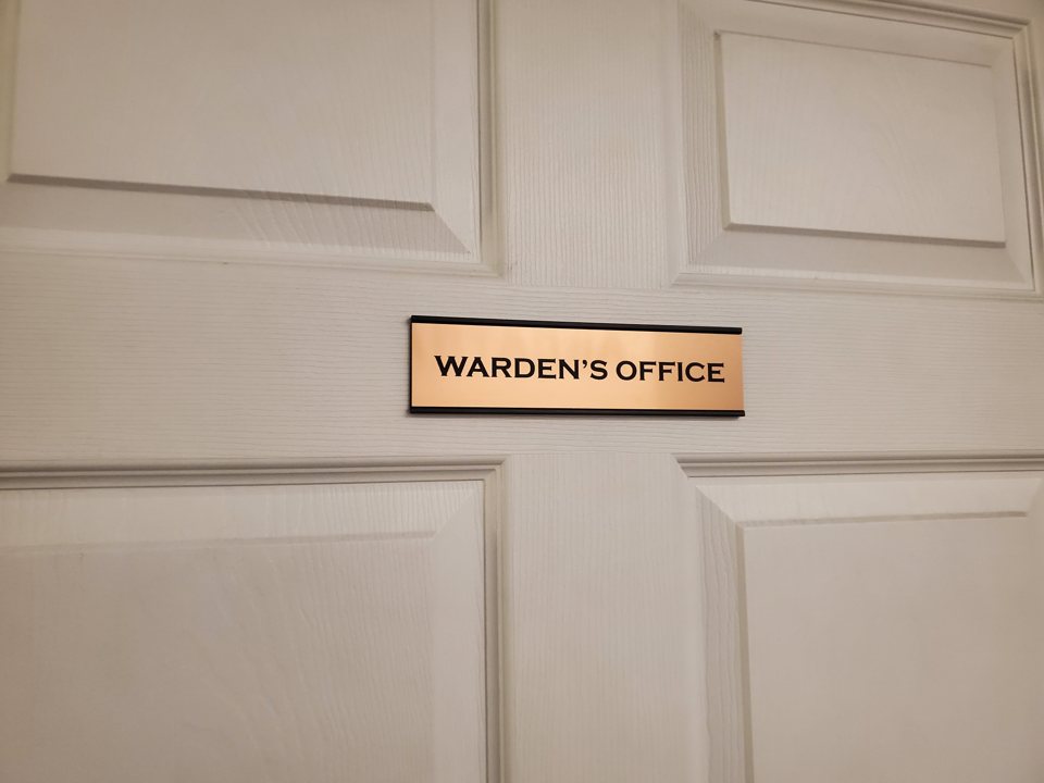 Fehér kazettás ajtó „Warden's Office” feliratú, rézszínű névtáblával