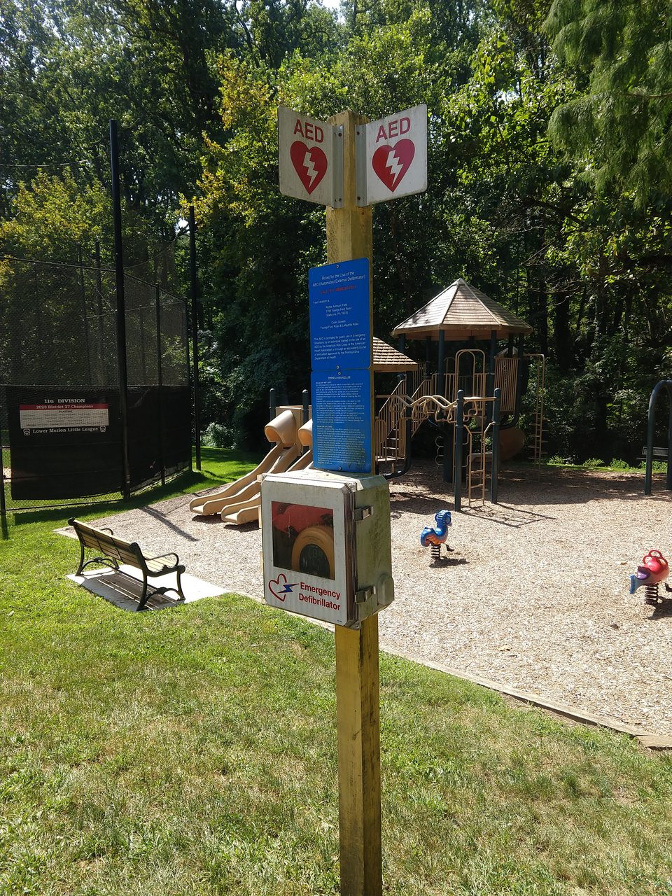 An outdoor AED box and red signs on a post next to a playground