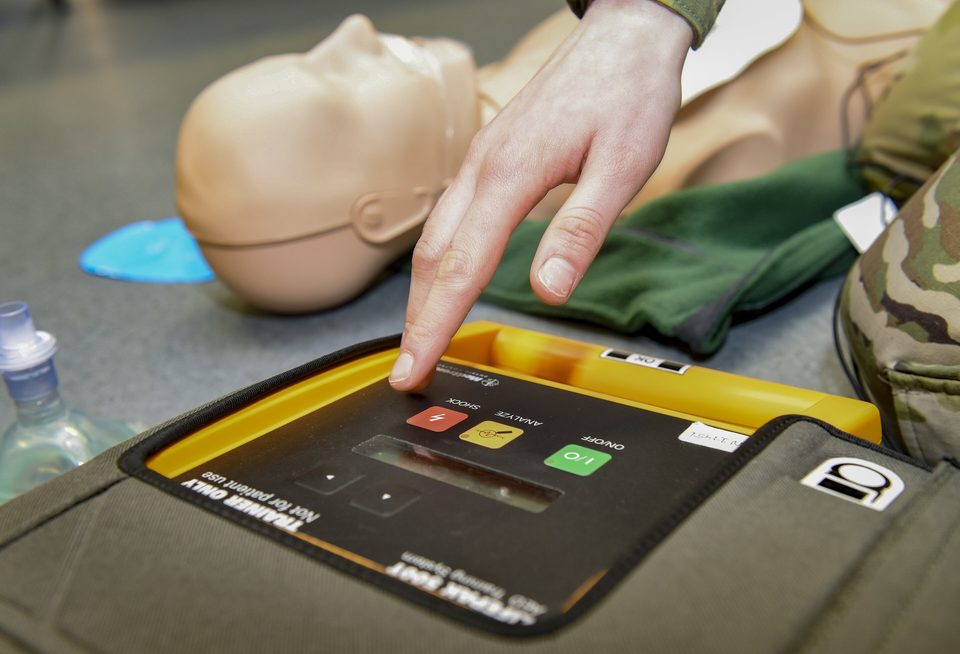 A finger presses the shock button on a training AED next to a CPR manikin