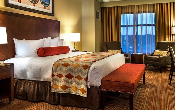 A guest room with a large bed, an orange bolster pillow and armchairs by the window