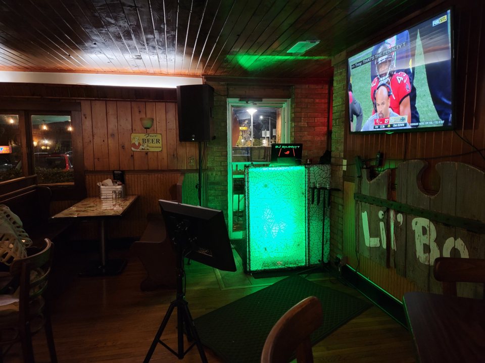 The karaoke corner with a green-lit DJ booth, speakers, a monitor and wood-paneled walls