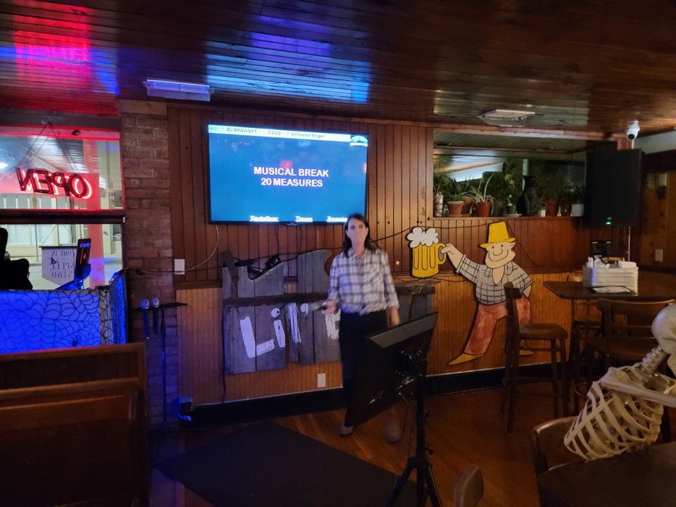 A wood-paneled bar room where a guest sings in the karaoke corner below a lyrics screen