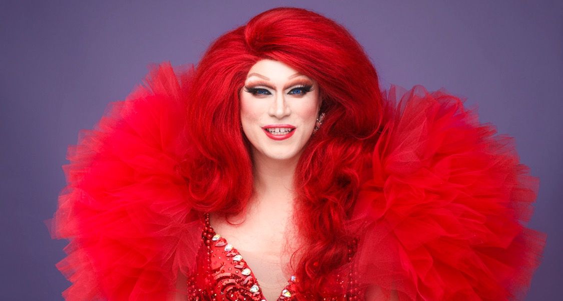 Drag performer in a red wig and voluminous red tulle collar against a purple backdrop
