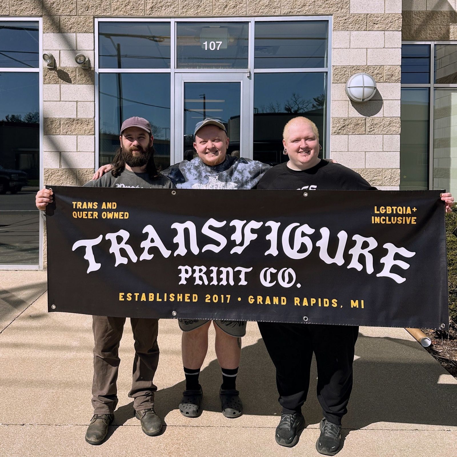 Three people hold a Transfigure Print Co. banner outside the shop