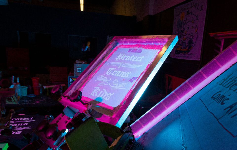 Screen-printing press with a “Protect Trans Kids” screen in pink light