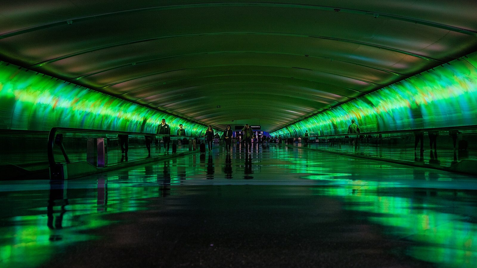 Travelers walking through the underground Light Tunnel between McNamara Terminal concourses, its curved walls glowing green and reflected on the polished floor.