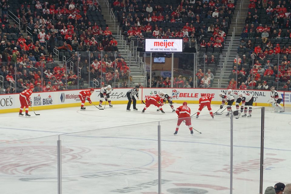 Faceoff between the Red Wings and the Arizona Coyotes at Little Caesars Arena, with red-clad fans behind the glass