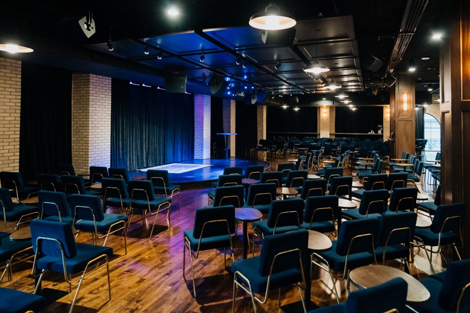 A River City Improv Midtown-terme kék kárpitos székekkel, kis asztalokkal és kék fényben álló színpaddal