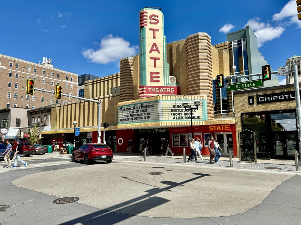 The State Theater's vertical STATE sign and marquee on a sunny day at the corner of State Street, with people walking past