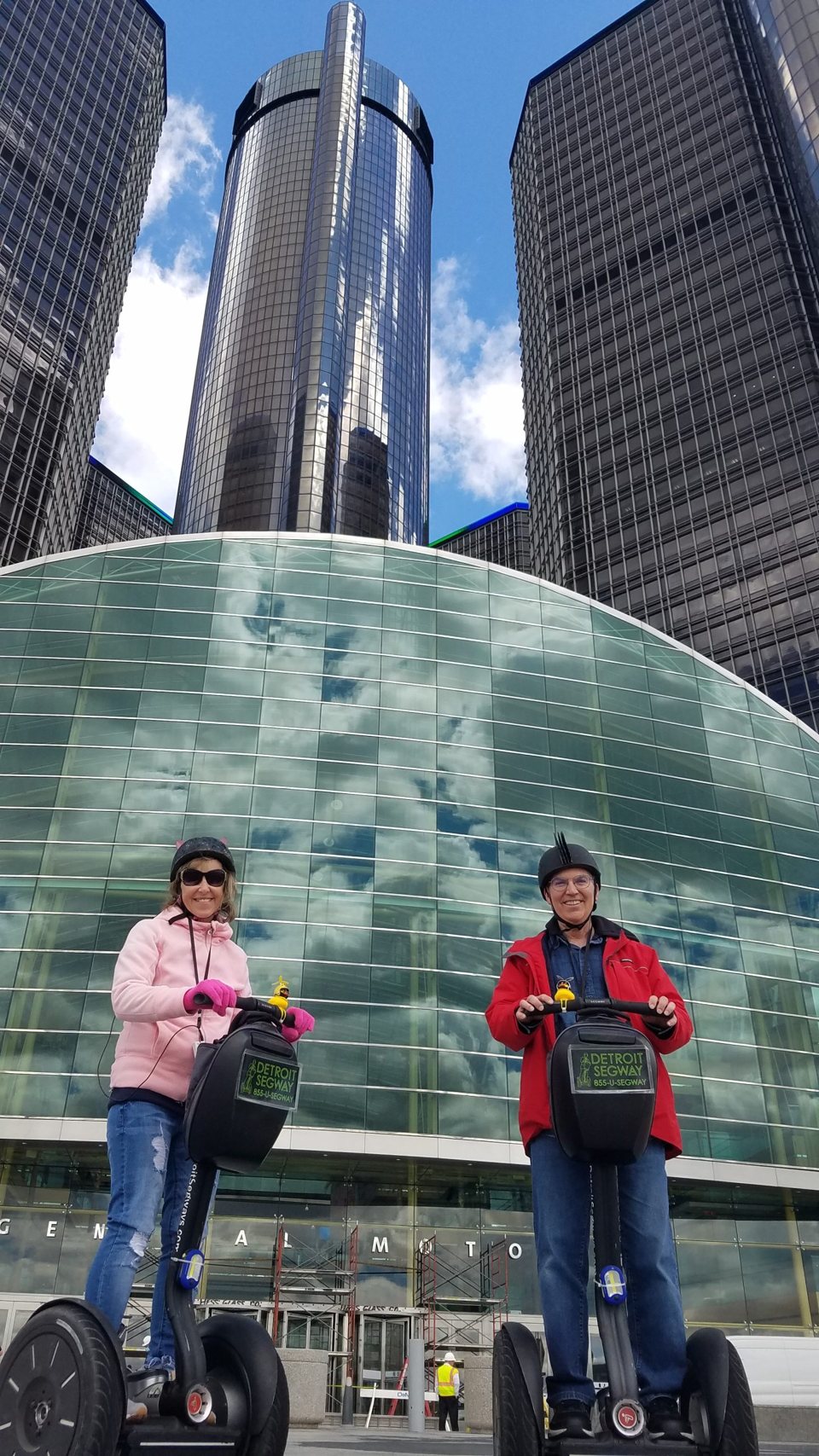Two riders on Segways stand in front of the Renaissance Center towers, with the company's logo on their bags